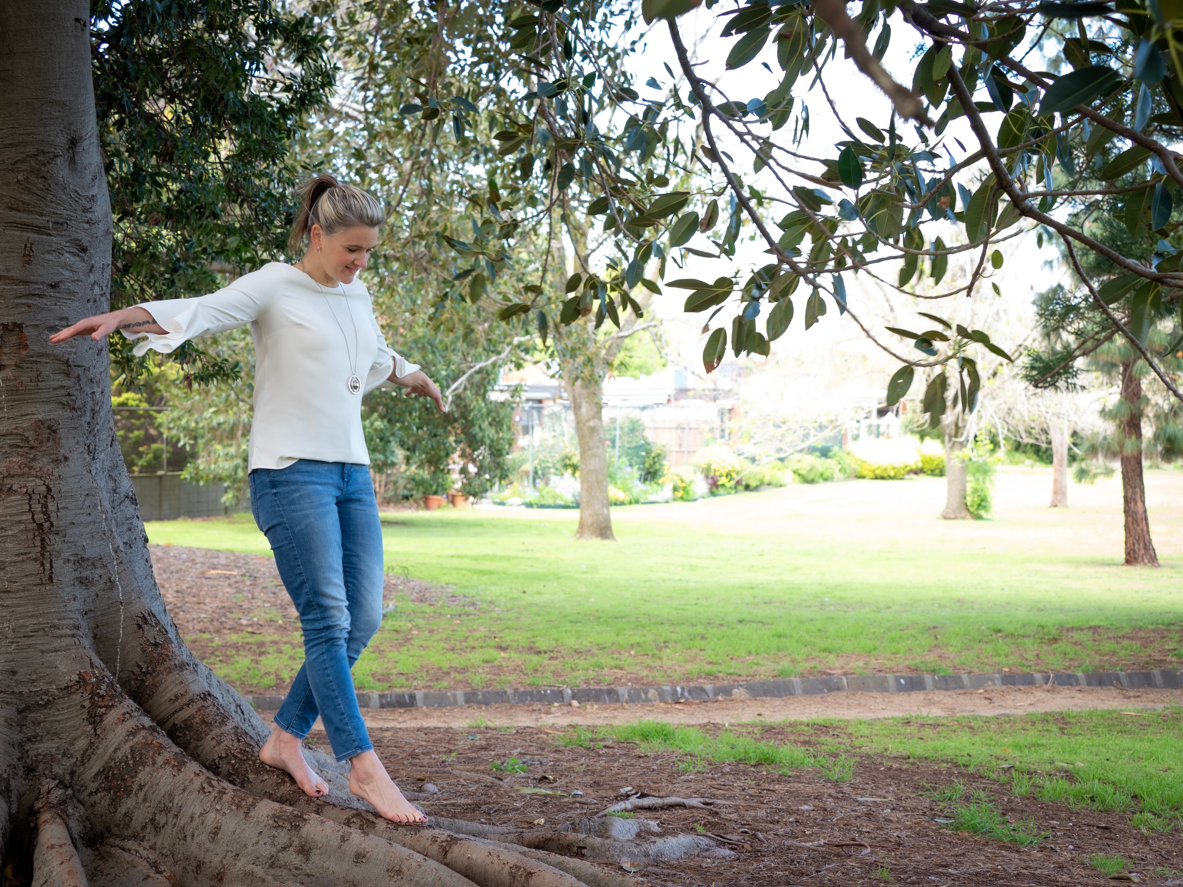 Linds, barefoot, balancing along the broad surface roots of a large fig tree in a park.